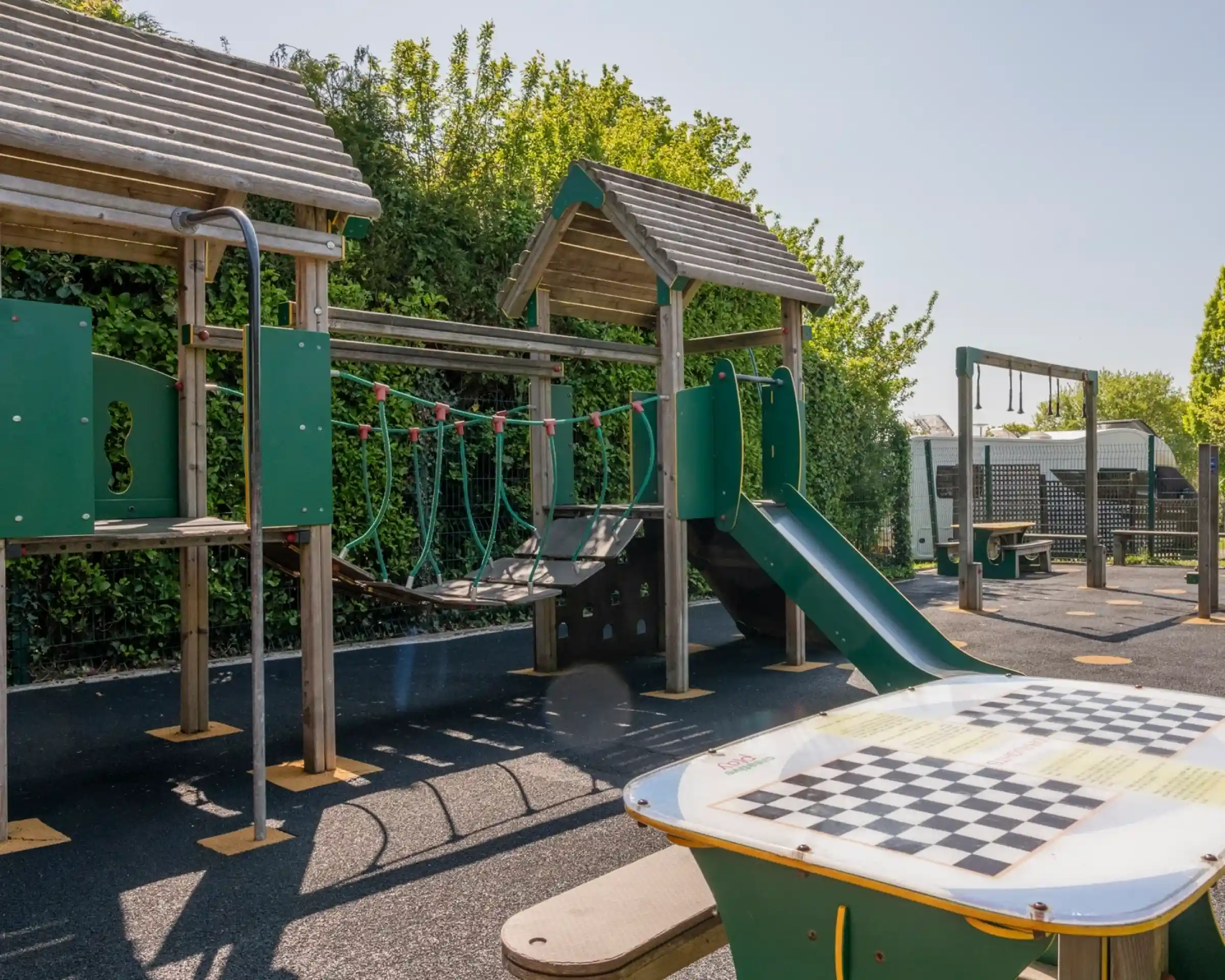 Playground with a green climbing structure featuring a slide and rope bridge. A checkers table is in the foreground, surrounded by trees and a sunny sky.