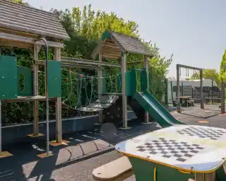 Playground with a green climbing structure featuring a slide and rope bridge. A checkers table is in the foreground, surrounded by trees and a sunny sky.