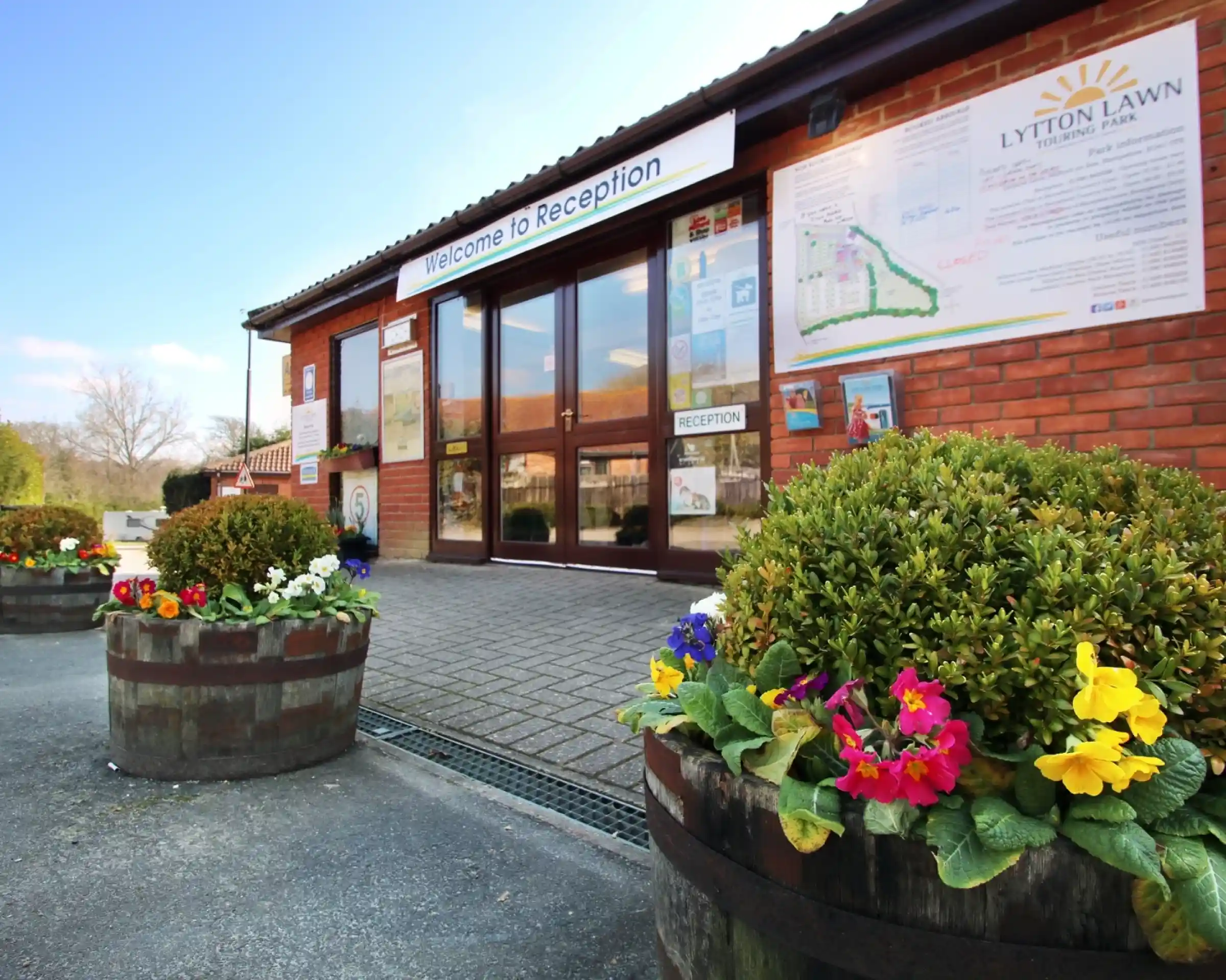 A welcoming reception area featuring large glass doors, with a sign that reads "Welcome to Reception." Colorful flower pots with blooming flowers are placed outside on a paved pathway. A map is displayed on the wall, along with various informational materials.