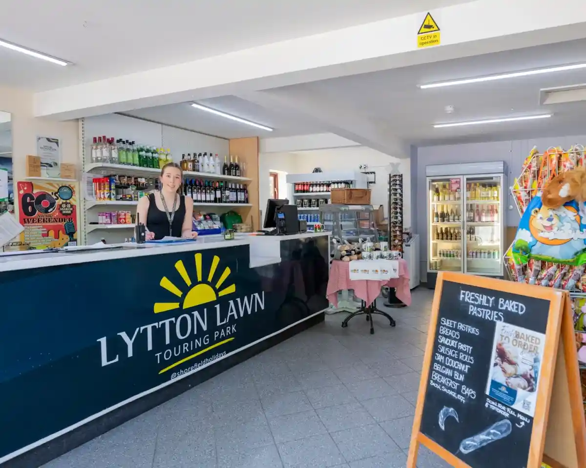 A welcoming reception area at Lytton Lawn Touring Park with a smiling staff member behind the counter. Shelves stocked with various drinks and snacks, alongside a display of freshly baked pastries and colorful candy items.