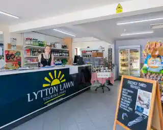 A welcoming reception area at Lytton Lawn Touring Park with a smiling staff member behind the counter. Shelves stocked with various drinks and snacks, alongside a display of freshly baked pastries and colorful candy items.