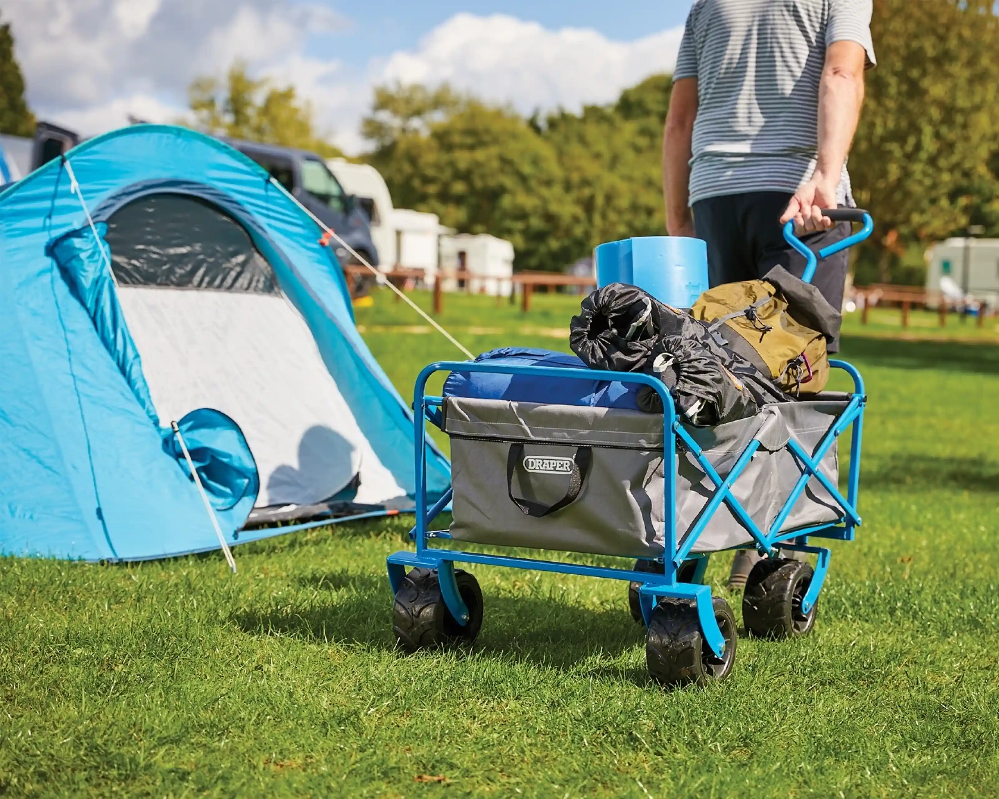 A person pulls a blue, wheeled cart filled with camping gear beside a blue tent in a grassy camping area. Trees and other tents are visible in the background under a partly cloudy sky.
