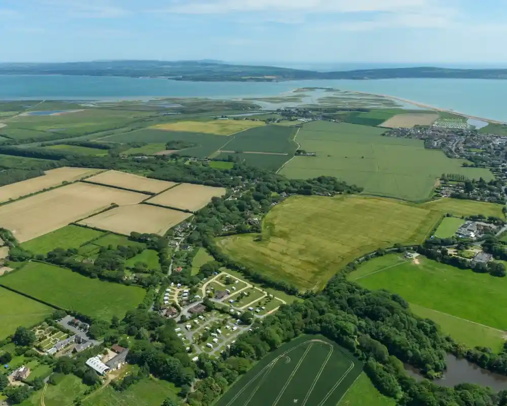 Aerial view of lush green fields, scattered trees, and a coastal area with blue waters. The landscape features patches of farmland, a river winding through, and residential areas in the distance.