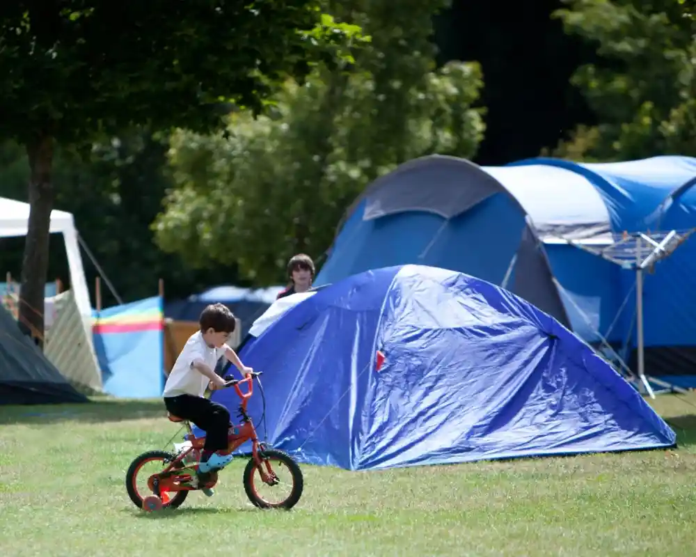 A young child rides a small bicycle in a campsite, with blue tents set up in the background surrounded by trees.