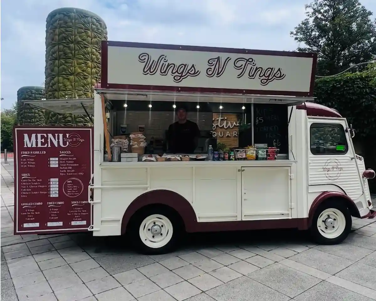 A retro-style food truck named "Wings N Tings" with a menu displayed prominently on the side. The truck is adorned with red and white colors, and a person stands behind the counter, ready to serve food in a vibrant outdoor setting. The surrounding area features greenery and paved stones.