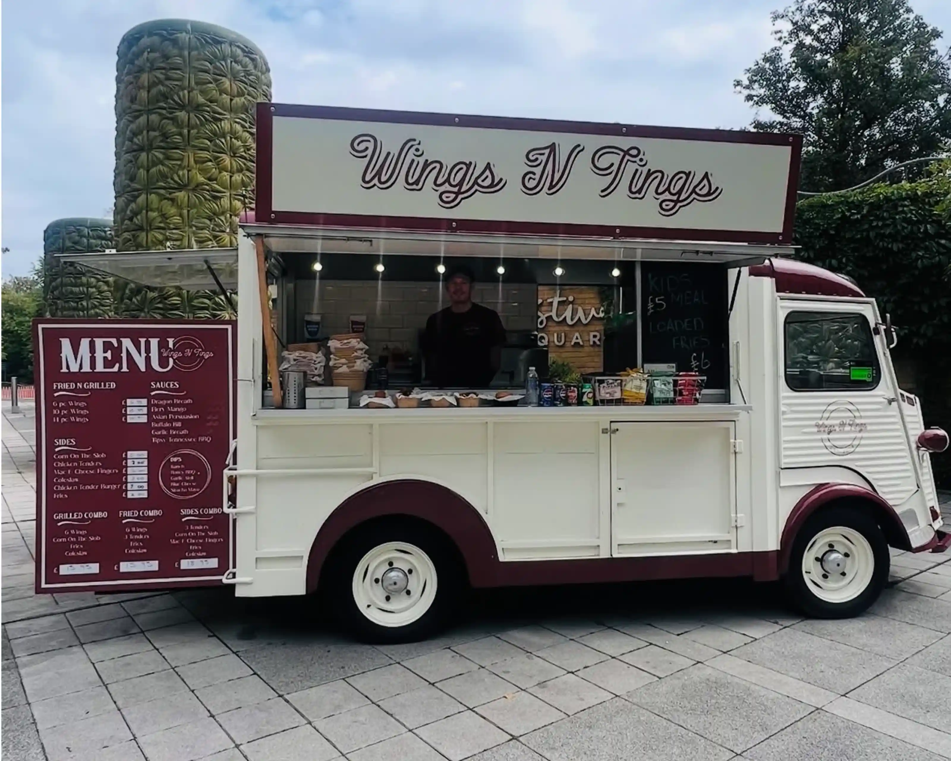 A retro-style food truck named "Wings N Tings" with a menu displayed prominently on the side. The truck is adorned with red and white colors, and a person stands behind the counter, ready to serve food in a vibrant outdoor setting. The surrounding area features greenery and paved stones.