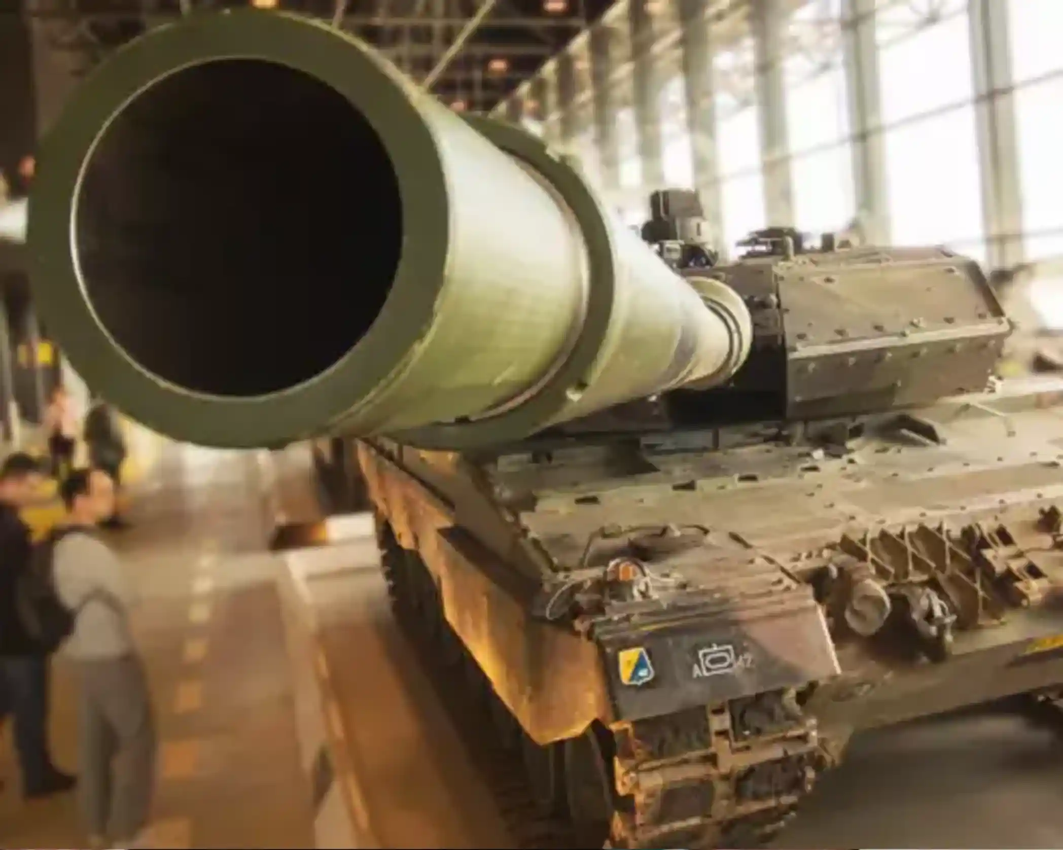 A large tank with a prominent cannon is displayed in an indoor exhibition space. People are gathered nearby, observing the tank's details. The environment features large windows that allow natural light to illuminate the scene.