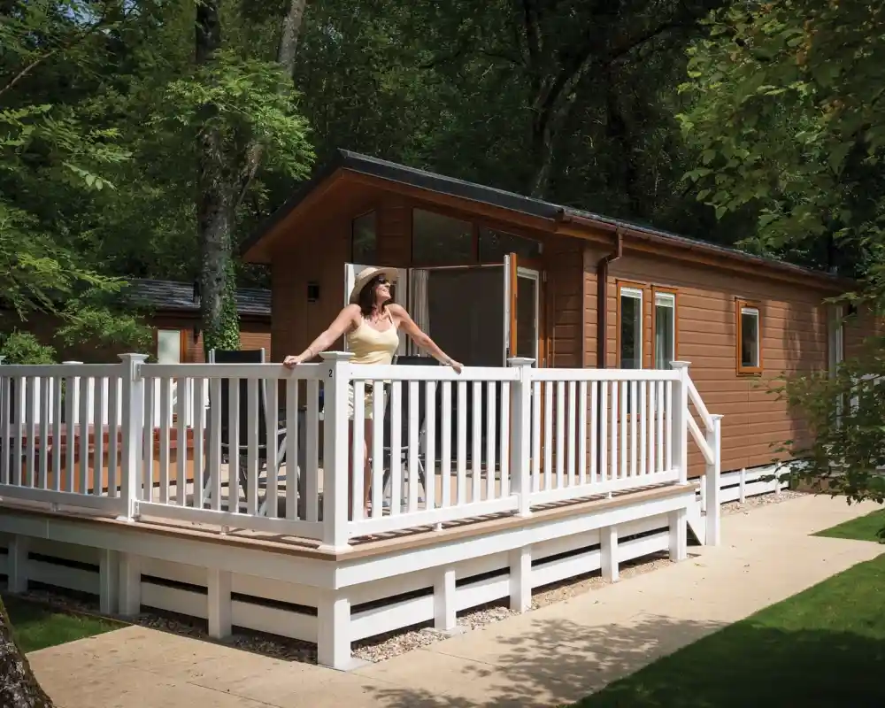 A woman in a light yellow dress stands on a wooden deck, enjoying the view of a brown cabin surrounded by greenery. The cabin features large windows and a sloping roof.