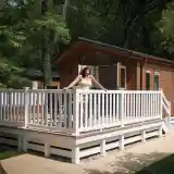 A woman in a light yellow dress stands on a wooden deck, enjoying the view of a brown cabin surrounded by greenery. The cabin features large windows and a sloping roof.