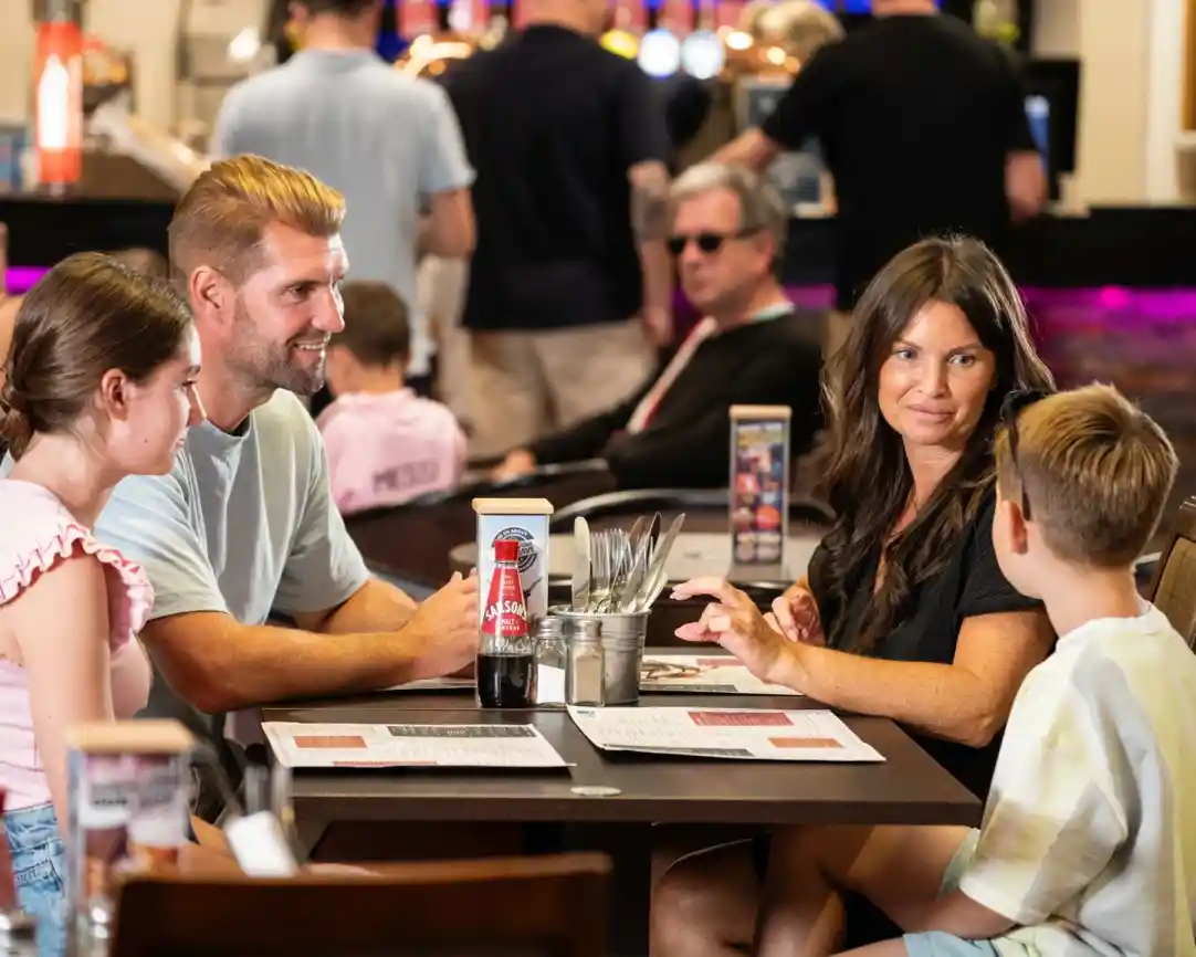 A family sits at a restaurant table engaged in conversation. Two children, a girl and a boy, are seated across from an adult couple. The atmosphere is lively, with other patrons in the background. Various condiments and menus are on the table.