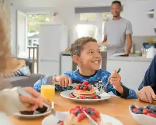 A young boy with curly hair smiles while holding a fork and sitting at a table with pancakes topped with berries. A glass of orange juice is in front of him. In the background, an adult male stands near the kitchen counter, smiling.