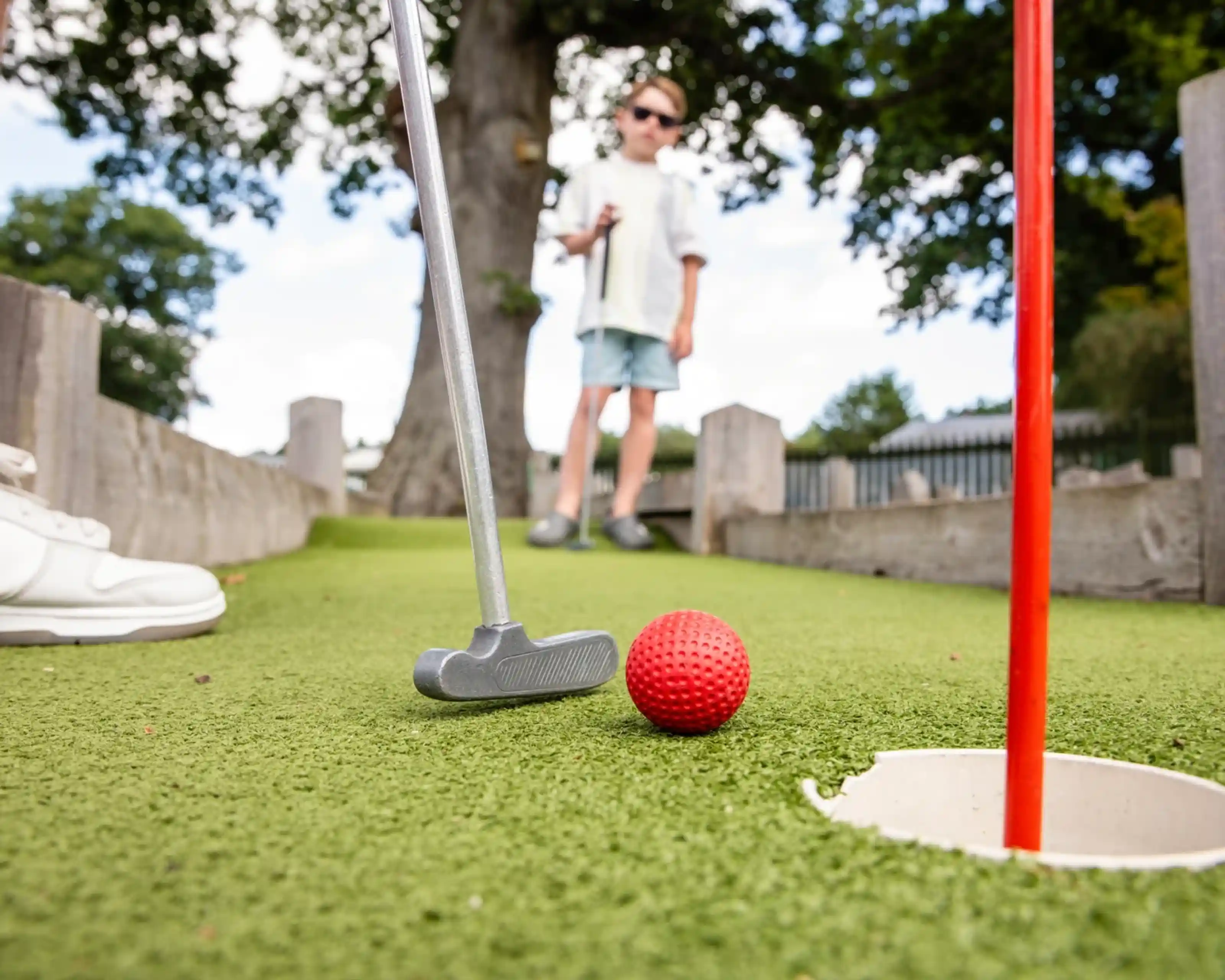 A close-up view of a mini-golf scene featuring a putter and a red golf ball near the hole. In the background, a child holding a golf club stands on the green. A large tree provides shade in the background.