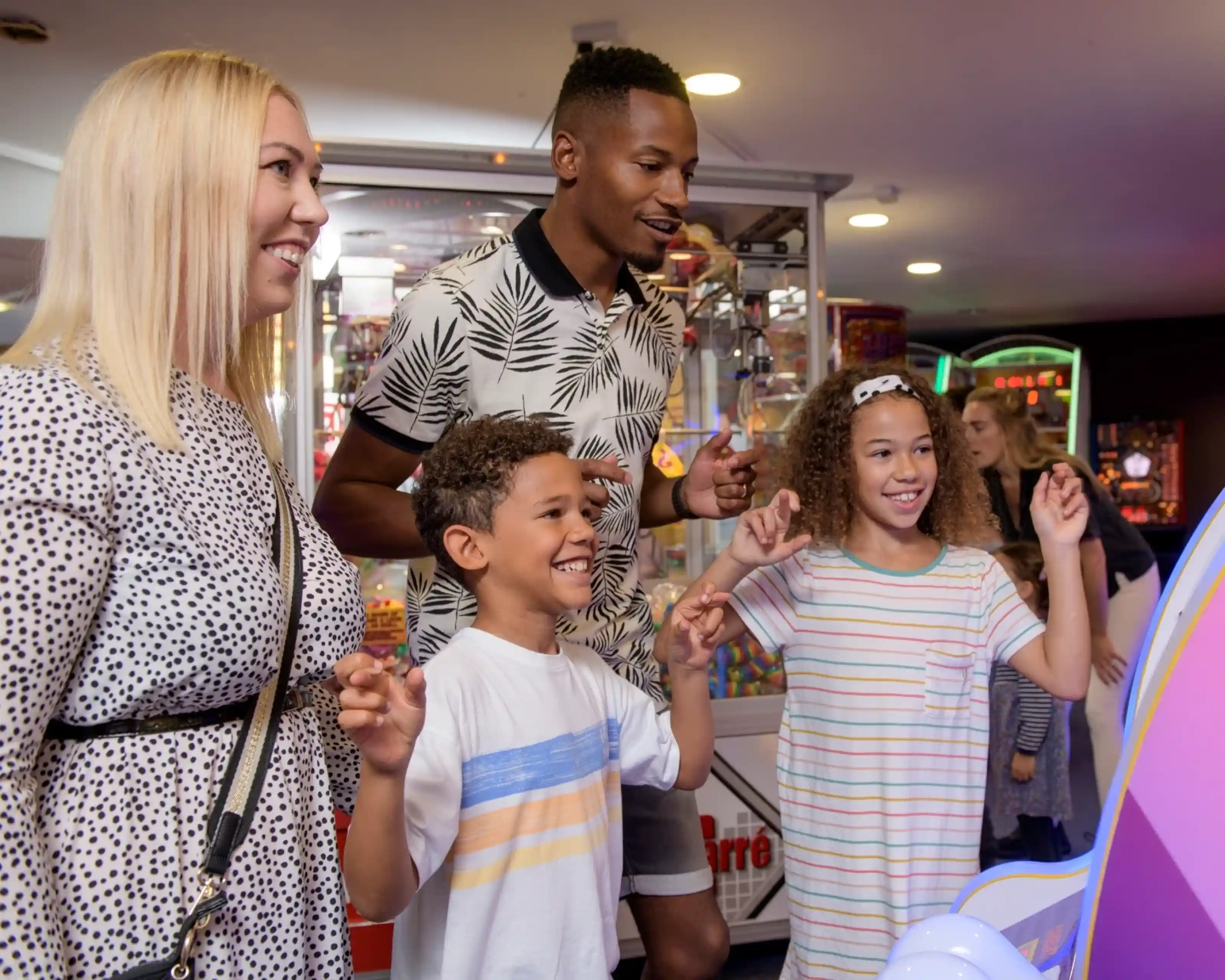 A family enjoys playing arcade games together, smiling and interacting with the game. A woman with blonde hair and a black-and-white printed dress stands next to a man in a patterned shirt. Two children, a boy and a girl, are engaged with the game, looking excited. The arcade is colorful with various games in the background.