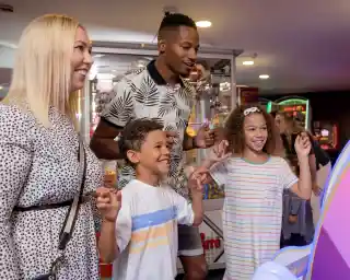 A family enjoys playing arcade games together, smiling and interacting with the game. A woman with blonde hair and a black-and-white printed dress stands next to a man in a patterned shirt. Two children, a boy and a girl, are engaged with the game, looking excited. The arcade is colorful with various games in the background.
