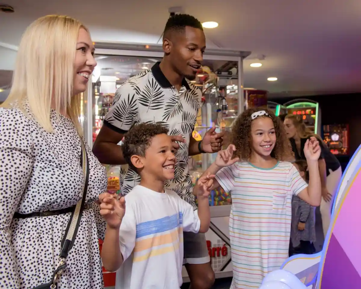 A family enjoys playing arcade games together, smiling and interacting with the game. A woman with blonde hair and a black-and-white printed dress stands next to a man in a patterned shirt. Two children, a boy and a girl, are engaged with the game, looking excited. The arcade is colorful with various games in the background.