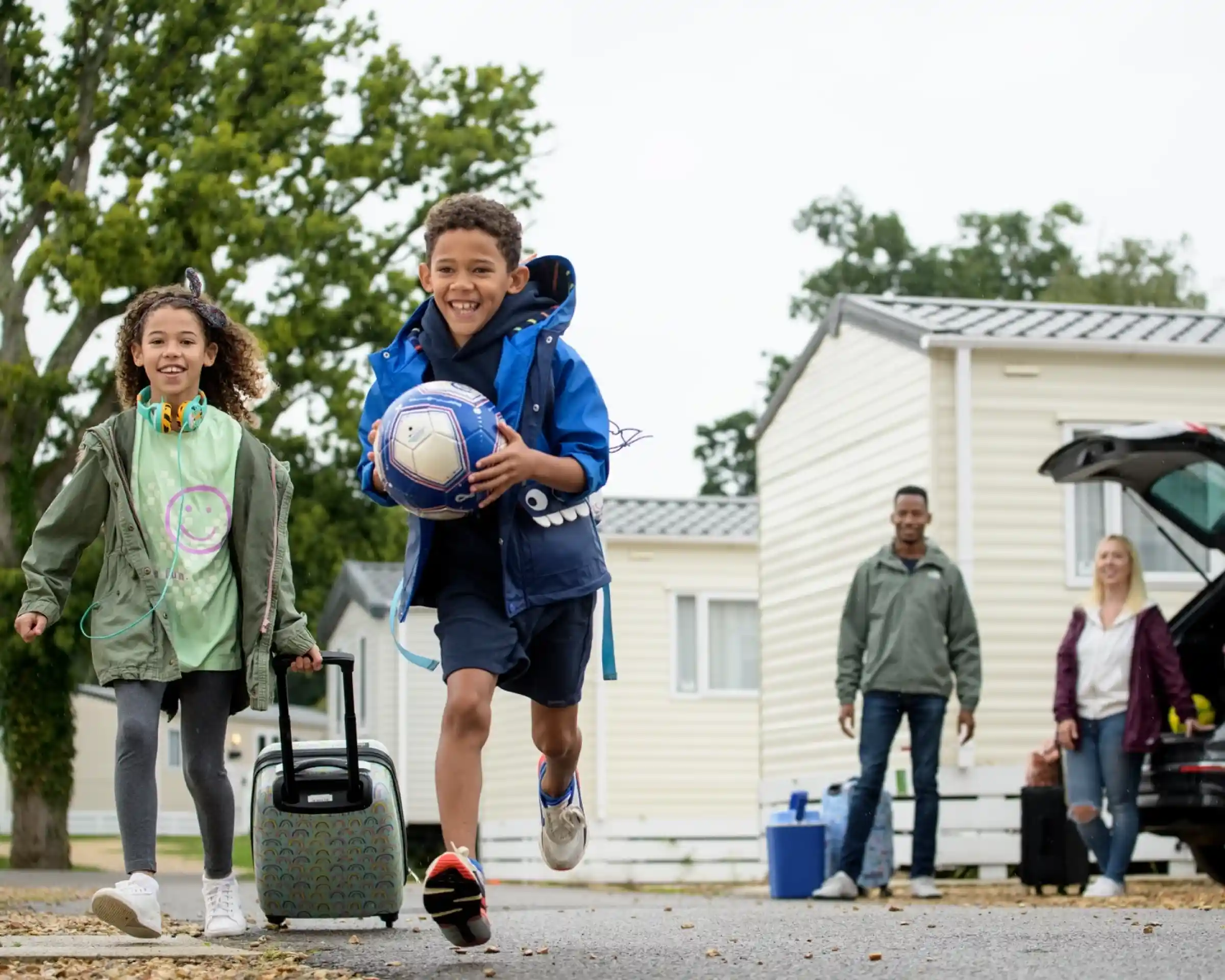 Two children, a boy with a soccer ball and a girl pulling a suitcase, run happily down a pathway in a holiday park. In the background, two adults stand by a car, smiling. Mobile homes and trees are visible around them.