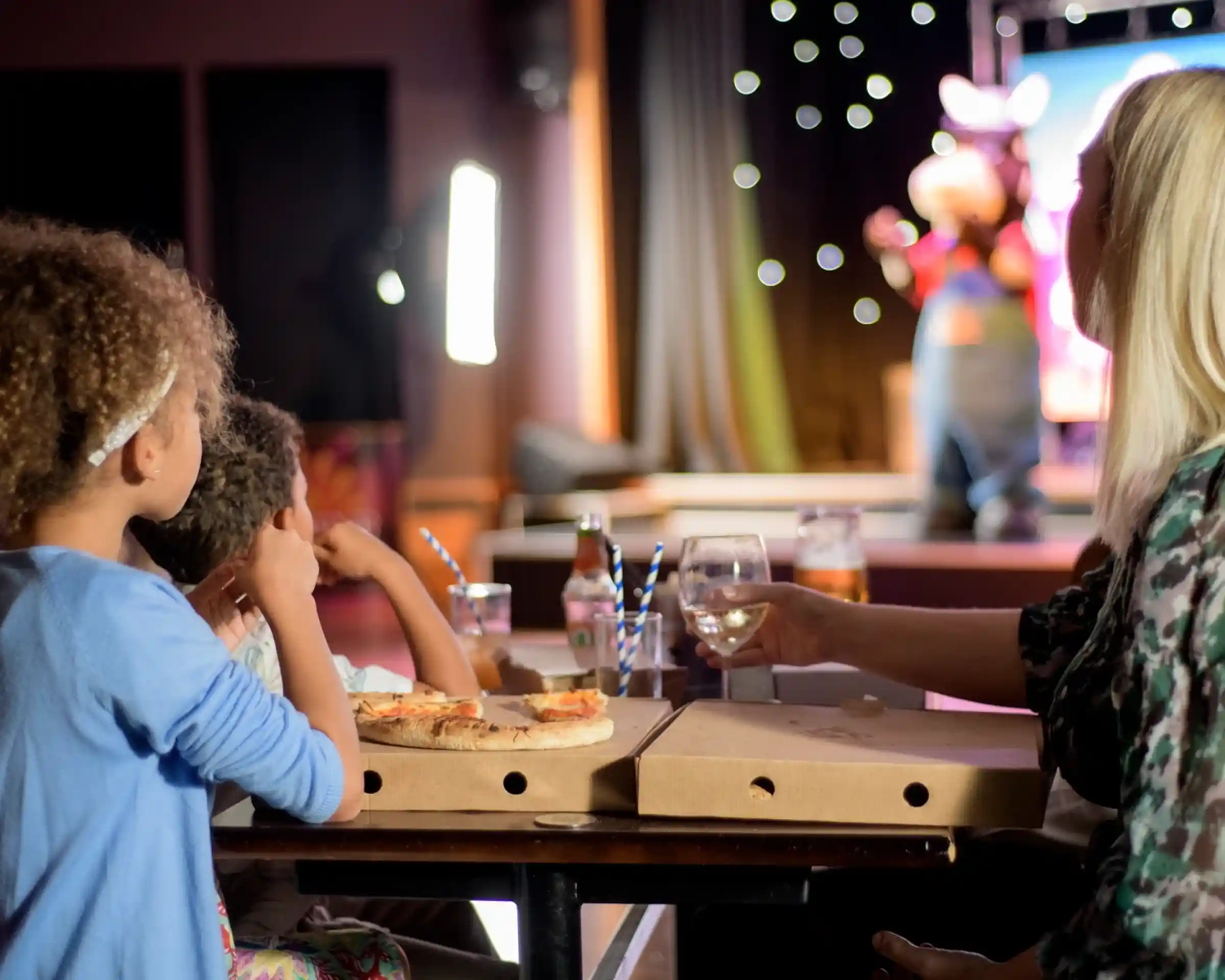 Two children and an adult sit at a table with pizza boxes, watching a performance on stage. The stage features a character in costume, with colorful lights and a festive atmosphere.