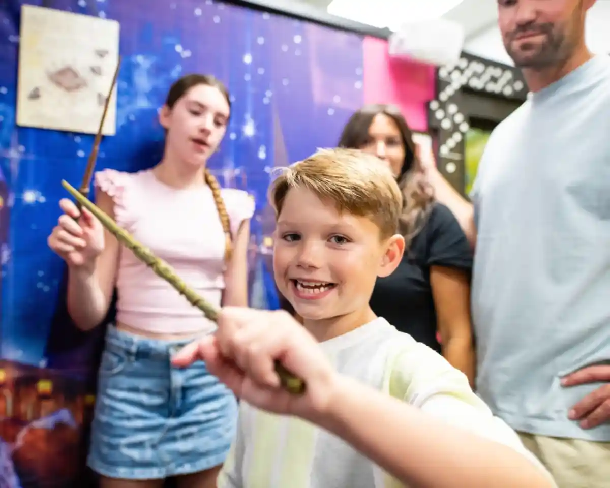A young boy smiles and holds a wand in the foreground, with two other people in the background. One is a girl who appears to be casting a spell, and another is a woman with dark hair. The setting features a colorful, themed backdrop.