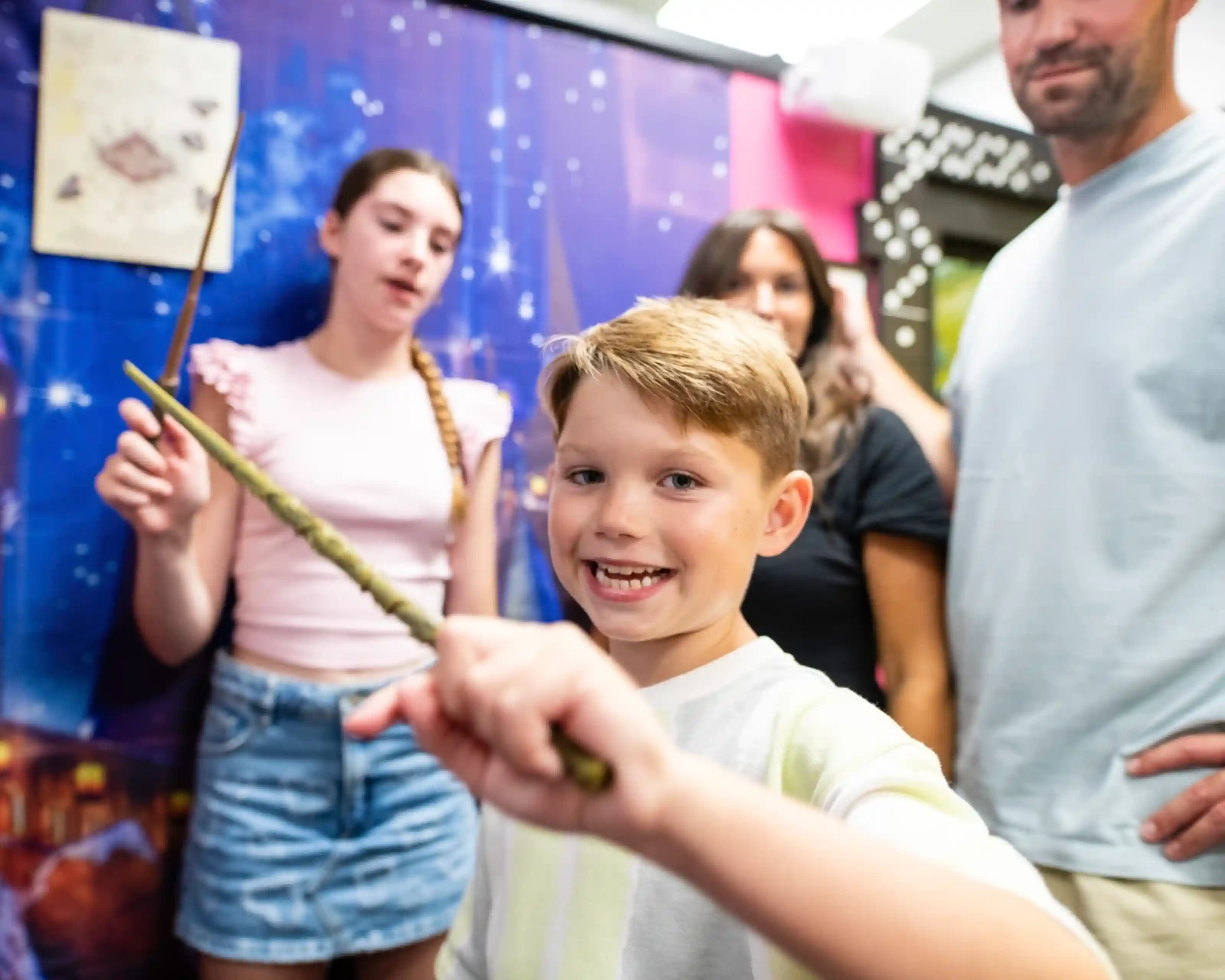 A young boy smiles and holds a wand in the foreground, with two other people in the background. One is a girl who appears to be casting a spell, and another is a woman with dark hair. The setting features a colorful, themed backdrop.