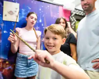 A young boy smiles and holds a wand in the foreground, with two other people in the background. One is a girl who appears to be casting a spell, and another is a woman with dark hair. The setting features a colorful, themed backdrop.