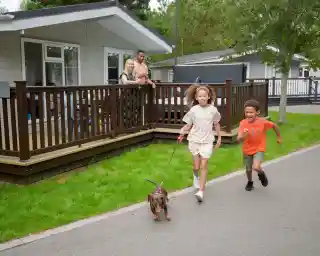 Two children run along a path outside a vacation cabin, holding a leash attached to a small dog. A man and a woman stand on the porch of the cabin, watching and smiling. The scene features green grass and trees in the background.