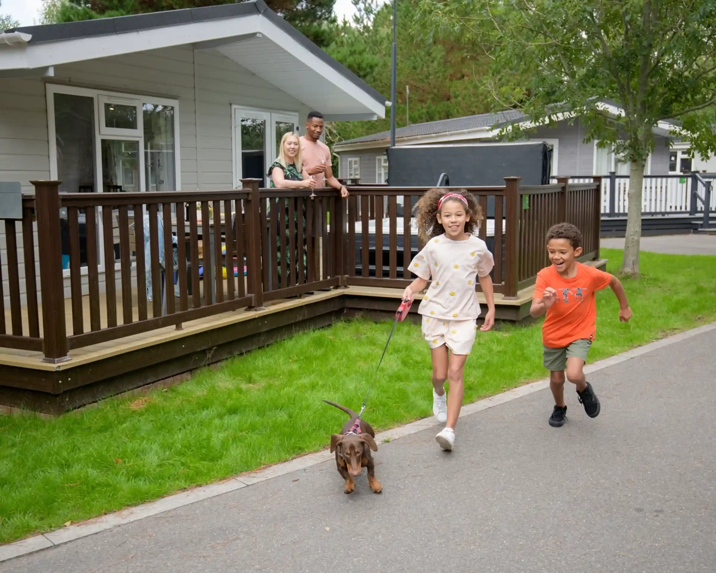 Two children run along a path outside a vacation cabin, holding a leash attached to a small dog. A man and a woman stand on the porch of the cabin, watching and smiling. The scene features green grass and trees in the background.