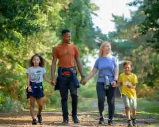 A family of four walks hand in hand along a wooded path. Two children, a girl and a boy, are on the left and right, respectively, wearing casual attire. The parents, a man and a woman, are in the center, both smiling. Lush green trees and sunlight create a warm, inviting atmosphere.