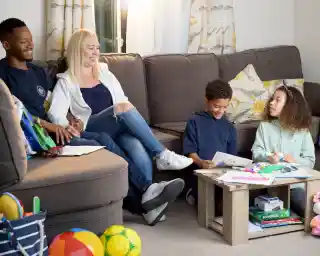 A family of four sits together in a cozy living room. An adult man and woman smile while seated on a couch, and two children are on the floor, engaged in a creative activity with coloring books and art supplies. Colorful toys are scattered around the room.