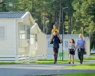 A man carries a boy on his shoulders while walking with a girl and a woman through a pathway lined with holiday cottages in a wooded area. Bright sunlight illuminates the scene, enhancing the cheerful atmosphere.