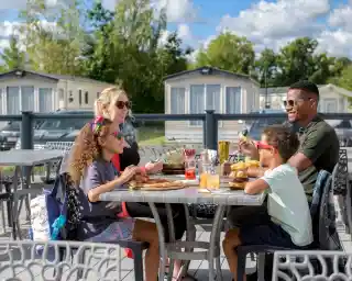 A cheerful family enjoys a meal at an outdoor table. The group includes two children wearing sunglasses and smiling, seated alongside an adult woman and a man. The table is set with food and drinks, with a background of mobile homes and a blue sky dotted with clouds.