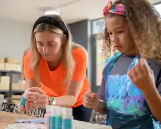 A woman in an orange shirt assists a girl with curly hair while they work on a craft project at a table. The girl is focused on her materials, and both are using colorful paint supplies in a bright, airy workshop setting.