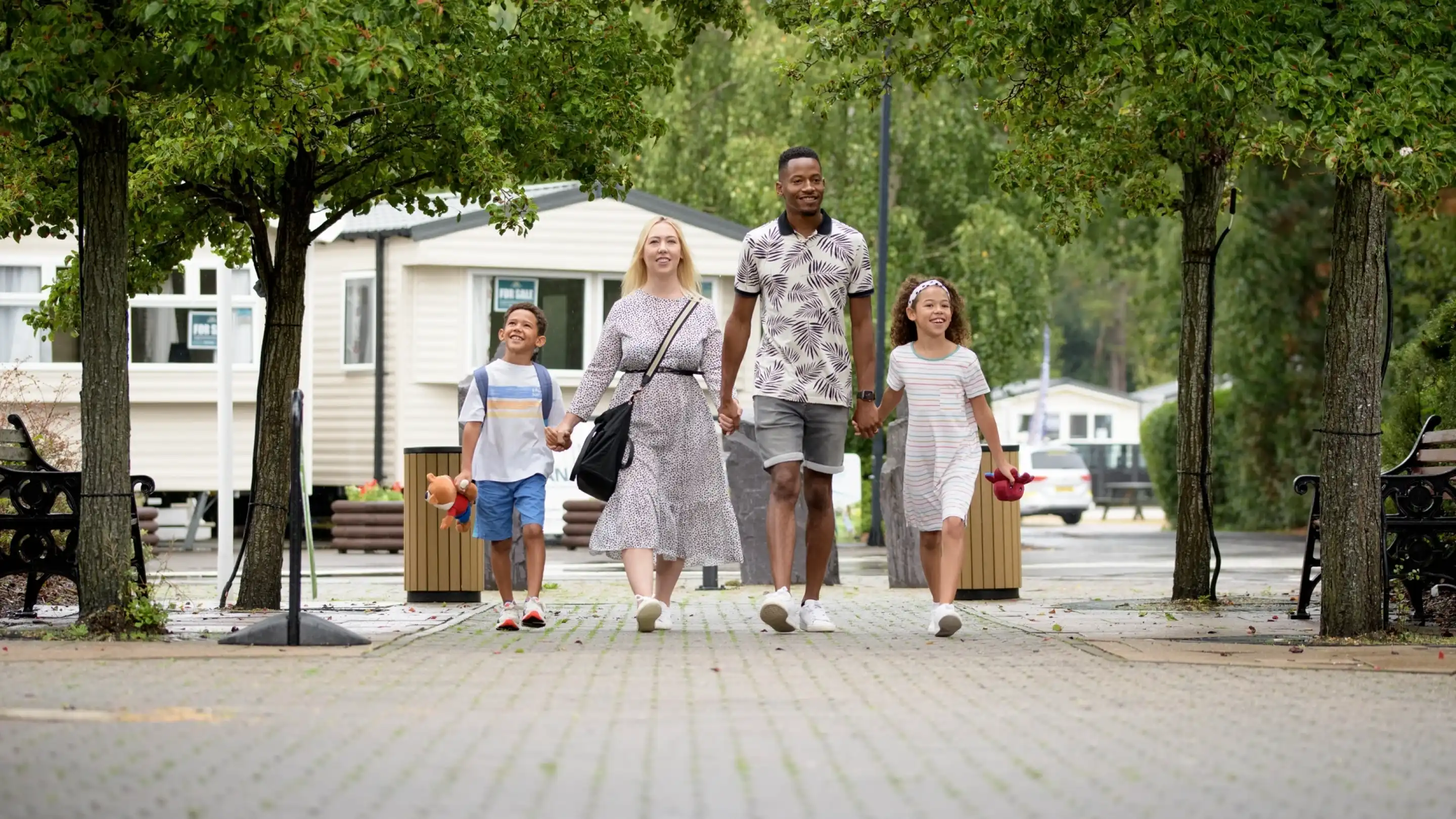 A family of four walks hand in hand along a tree-lined pathway. A woman in a flowing dress holds hands with two children, a boy and a girl, while a man walks beside them. They are smiling and enjoying their time together.