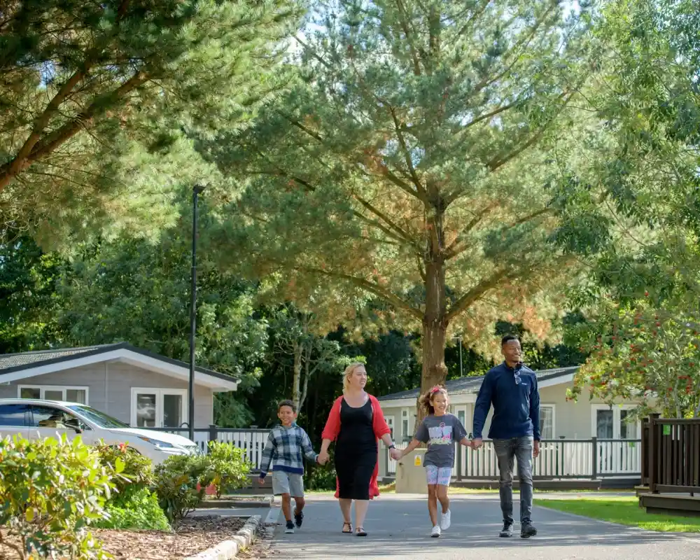 A woman and a man walk hand in hand with two children along a pathway lined with trees and residential buildings. The sun is shining, creating a warm and inviting atmosphere.