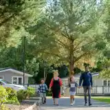 A woman and a man walk hand in hand with two children along a pathway lined with trees and residential buildings. The sun is shining, creating a warm and inviting atmosphere.