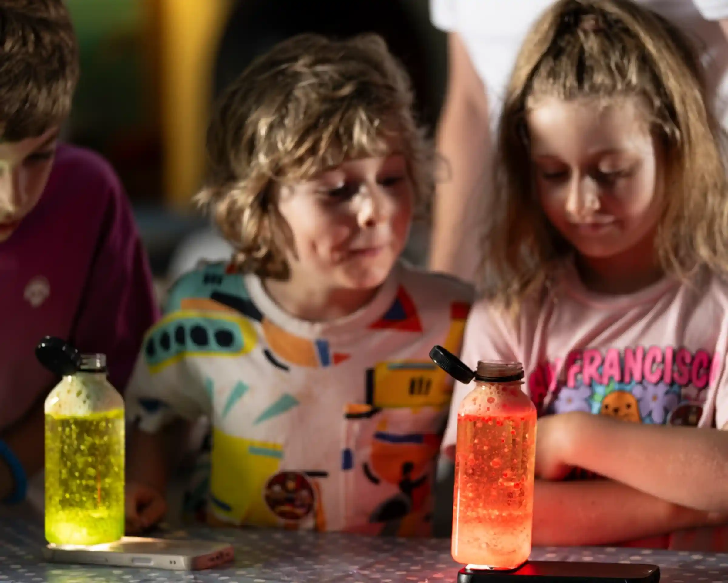 Children gather around glowing bottles filled with colorful liquids, one yellow and one red. They display expressions of wonder and curiosity as they observe the bottles.