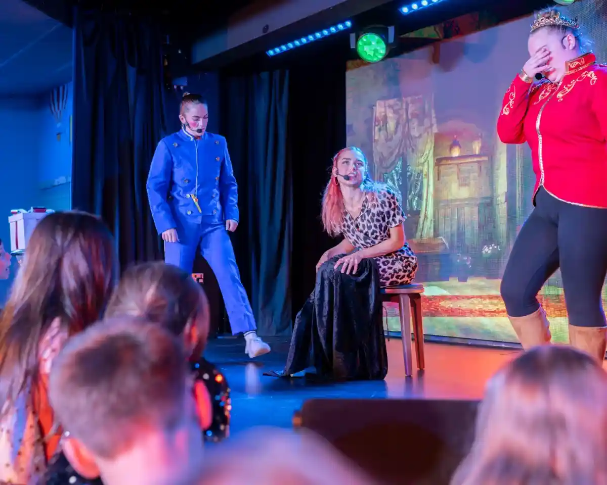 A theatrical performance featuring three actors on stage. One actor in a blue uniform stands on the left, while another in a red costume laughs with their head down on the right. The central actor, dressed in a leopard-print outfit and black skirt, sits on a stool, looking at the others. Children in the audience are visible in the foreground, watching intently. Colorful stage lighting enhances the vibrant atmosphere.