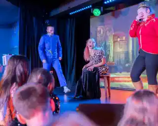 A theatrical performance featuring three actors on stage. One actor in a blue uniform stands on the left, while another in a red costume laughs with their head down on the right. The central actor, dressed in a leopard-print outfit and black skirt, sits on a stool, looking at the others. Children in the audience are visible in the foreground, watching intently. Colorful stage lighting enhances the vibrant atmosphere.