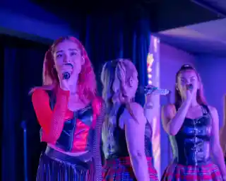 A singer with red hair and a black and red outfit performs on stage, holding a microphone. Two other female performers, dressed in matching outfits, sing in the background. Colorful stage lights illuminate the scene.