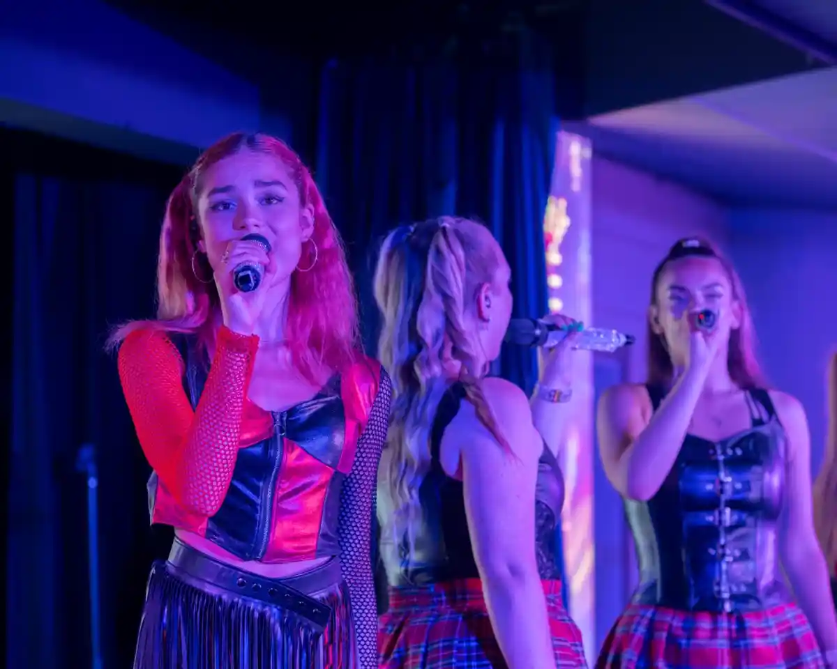 A singer with red hair and a black and red outfit performs on stage, holding a microphone. Two other female performers, dressed in matching outfits, sing in the background. Colorful stage lights illuminate the scene.