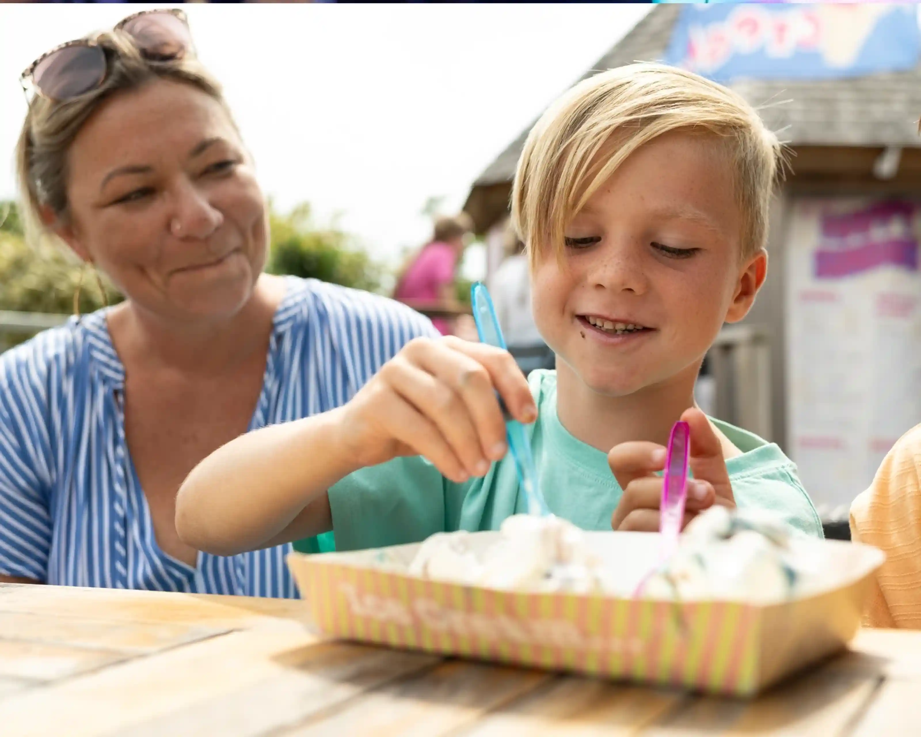 A child with blonde hair enjoys ice cream, using a colorful spoon, while a woman with light brown hair smiles at him, sitting outdoors at a wooden table.