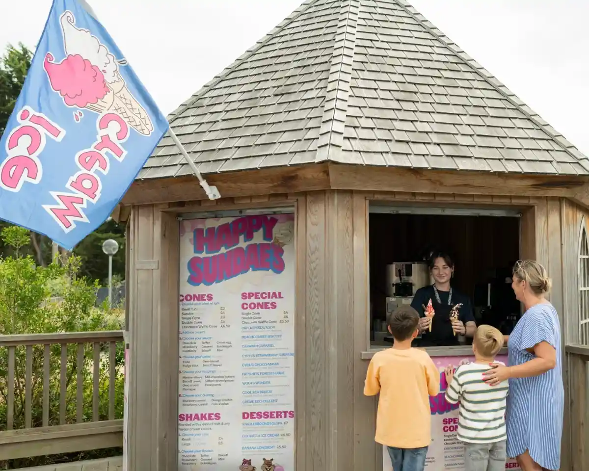 A woman and two children stand in front of a wooden ice cream stand with a shingled roof. The stand features a colorful menu displaying various ice cream options. A staff member serves ice cream cones to the customers. A flag with "Ice Cream" is flying above the stand.