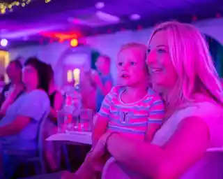 A mother and her young daughter smile joyfully while watching a performance in a colorful, dimly lit venue. The atmosphere is vibrant with purple and pink lighting, and other audience members can be seen in the background.
