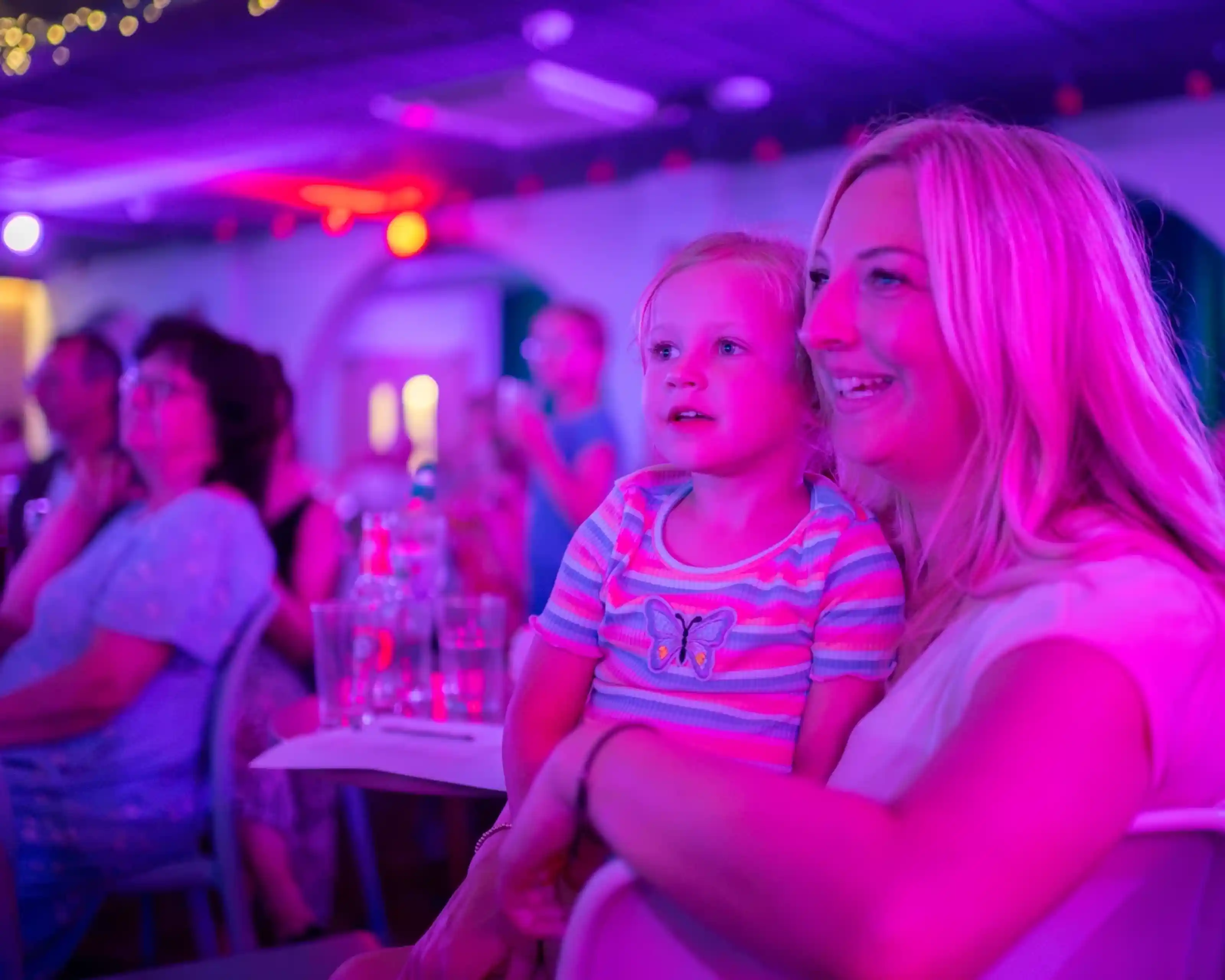 A mother and her young daughter smile joyfully while watching a performance in a colorful, dimly lit venue. The atmosphere is vibrant with purple and pink lighting, and other audience members can be seen in the background.