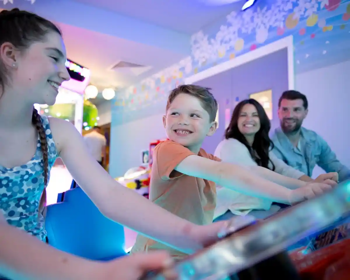 A smiling young boy plays a game at an arcade alongside a girl and two adults. They're enjoying their time together in a colorful, lively environment.