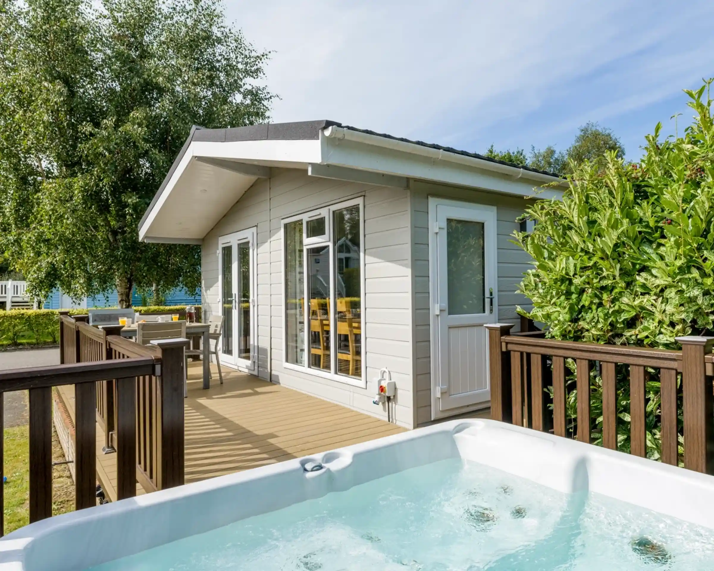 A stylish modern cabin with large windows, a wooden deck, and a hot tub in front. Surrounding greenery provides a serene atmosphere.