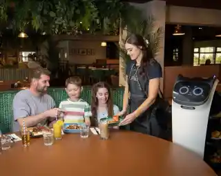 A family sits at a table in a restaurant, enjoying a meal. A waiter interacts with them while a service robot, equipped with a screen and wheels, delivers food. The setting features lush greenery and a cozy atmosphere.