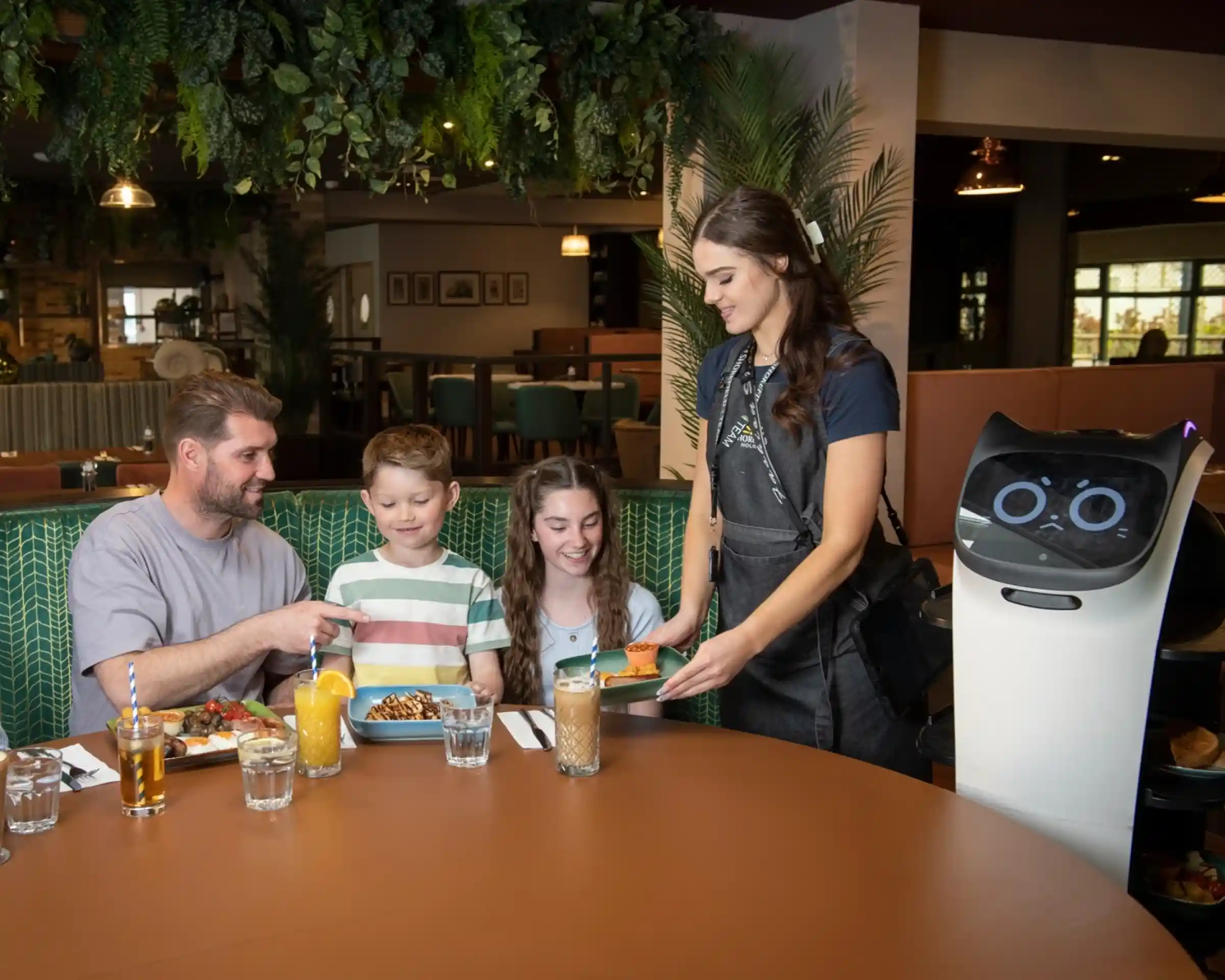 A family sits at a table in a restaurant, enjoying a meal. A waiter interacts with them while a service robot, equipped with a screen and wheels, delivers food. The setting features lush greenery and a cozy atmosphere.
