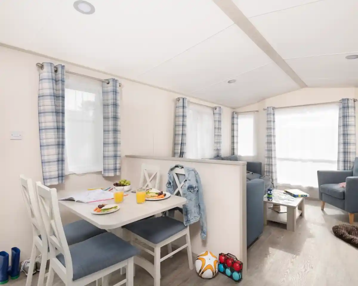 Modern interior of a cozy holiday home featuring a small dining table with four chairs, plates of food, and drinks. A blue sofa set and an armchair create a comfortable living space. Bright natural light flows through the windows, adorned with checkered curtains. A few colorful items, including a soccer ball and toys, are placed on the floor.