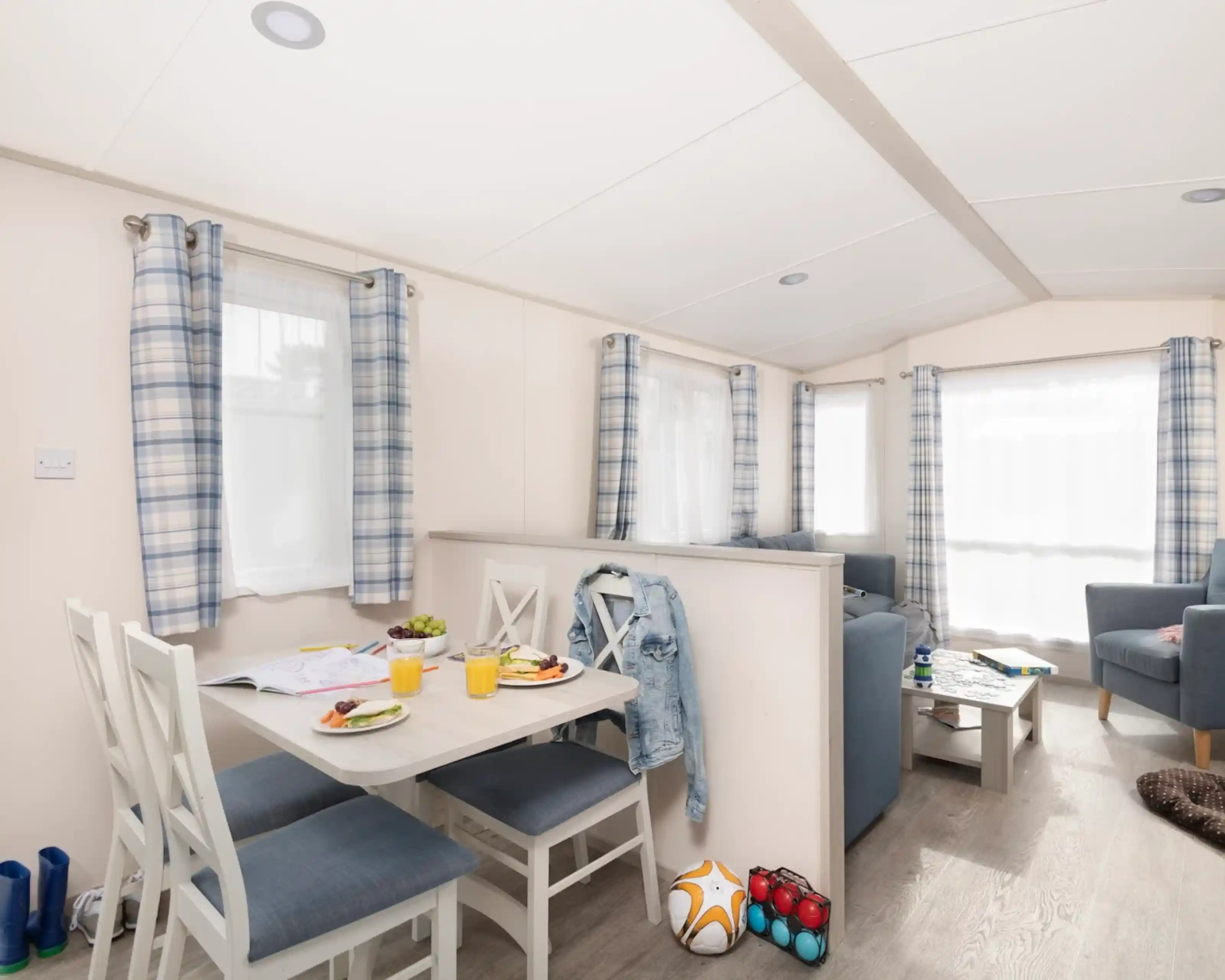 Modern interior of a cozy holiday home featuring a small dining table with four chairs, plates of food, and drinks. A blue sofa set and an armchair create a comfortable living space. Bright natural light flows through the windows, adorned with checkered curtains. A few colorful items, including a soccer ball and toys, are placed on the floor.