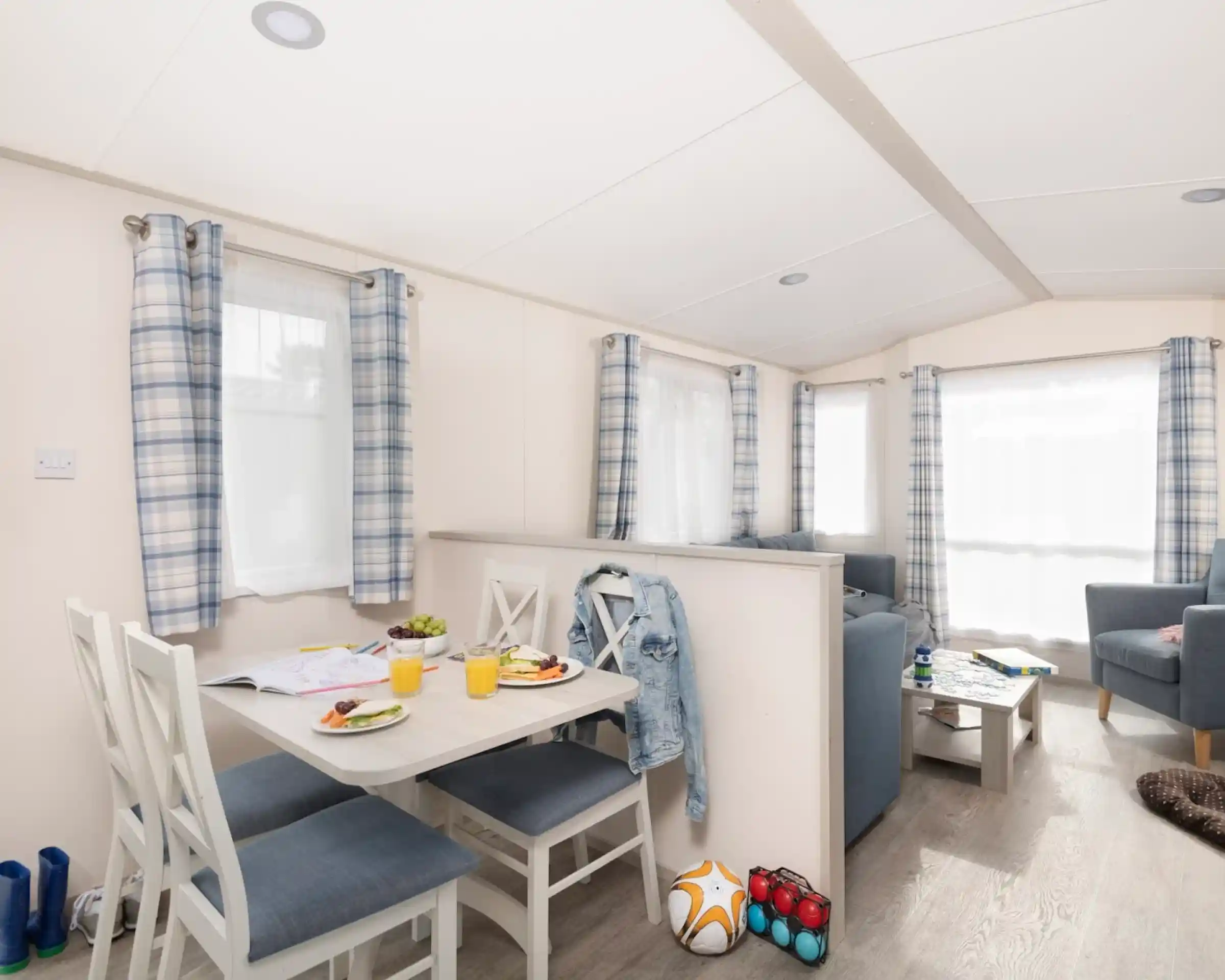 Modern interior of a cozy holiday home featuring a small dining table with four chairs, plates of food, and drinks. A blue sofa set and an armchair create a comfortable living space. Bright natural light flows through the windows, adorned with checkered curtains. A few colorful items, including a soccer ball and toys, are placed on the floor.